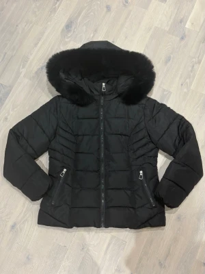 Svart pufferjacka med fuskpäls - Snygg svart pufferjacka med stor huva och mjuk fuskpäls runt kanten. Jackan har två dragkedjeförsedda fickor framtill och quiltade detaljer längs sidorna för en schysst passform. Perfekt för kalla vinterdagar och riktigt varm. Tyvärr har en knapp på luvan åkt av