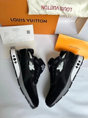 Louis Vuitton svarta sneakers - Snygga svarta sneakers från Louis Vuitton med vita detaljer och logga på sidan samt plös. Skorna har en mix av mocka och meshmaterial, svarta skosnören och en grå sula med ikoniska LV-symboler. Perfekta för dig som vill ha en exklusiv och trendig look.