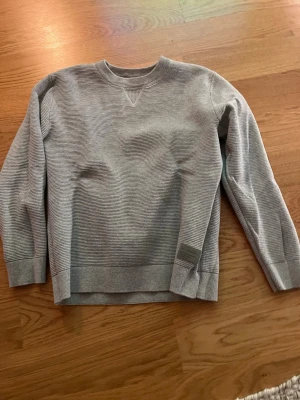 Grå ribbad sweatshirt - En stilren grå sweatshirt med diskret ribbad struktur och rund hals. Tröjan har långa ärmar, breda muddar och en liten patchdetalj vid ärmslutet. Perfekt för en avslappnad och enkel look.