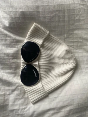 Vit ribbad mössa från C.P. Company - Säljer en vit ribbstickad mössa från C.P. Company med de ikoniska svarta glasögonlinserna framtill. Mössan är tillverkad i mjukt material och har en uppvikt kant som ger en snygg streetwear-look.