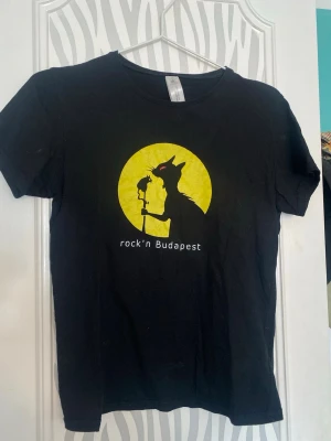Svart t-shirt Rock'n Budapest kattmotiv - Cool svart t-shirt från Rock'n Budapest med tryck av en katt i siluett mot en gul cirkel och texten 'rock'n Budapest' på bröstet. Perfekt för dig som gillar unika och grafiska motiv. Står M skulle säga att den passar mer som S