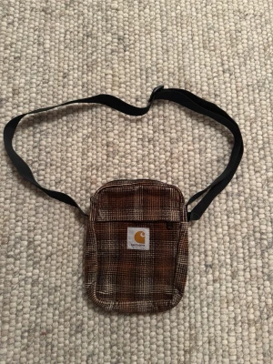 Carhartt Väska - Säljer en liten axelväska från Carhartt i brunt och beige rutigt tyg. Väskan har justerbar svart axelrem och en praktisk ficka framtill med Carhartt-logga. Perfekt för att bära det viktigaste och ger en cool streetwear-vibe.