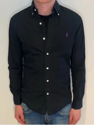 Svart skjorta från Ralph Lauren - Klassisk svart skjorta från Ralph Lauren med lila broderad logga på bröstet. Skjortan har knappar framtill och på kragen samt långa ärmar. Tillverkad i bomull och har en normal passform. Perfekt till jeans eller chinos för en clean look.