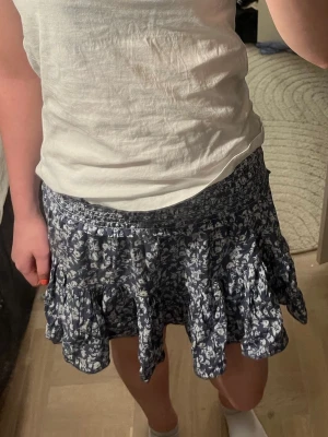 Blommig blå volangkjol - Supersöt kort kjol i blått med vitt blommönster och volangdetaljer. Perfekt till en varm sommar dag. Resår i midjan för extra komfort och snygg passform. Säljer den då den inte längre kommer till användning, kontakta mig om ni har några frågor💕