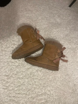 Bruna uggsliknande skor med rosetter  - Mysiga bruna stövlar  med fluffig kant och två söta rosetter bak. Utsidan är i mocka och insidan är fodrad med mjukt foder. Perfekta för kalla dagar och ger en riktigt cozy vibe.