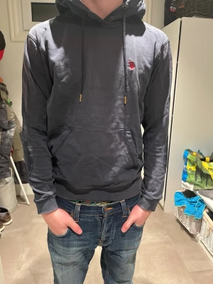 Mörkblå hoodie från Polo Ralph Lauren - Snygg mörkblå hoodie från Polo Ralph Lauren med röd broderad logga på bröstet och förstärkta armbågslappar. Klassisk känguruficka framtill och justerbar huva med mörkblåa  snören och gulddetaljer. Perfekt för en avslappnad och stilren look.