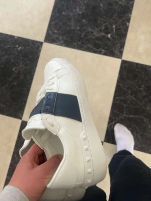 Vita sneakers med blå detalj Valentino - Säljer ett par vita sneakers från Valentino med bred mörkblå rem över sidan och nitar på sulan. Skorna har vita snören och är tillverkade i slätt läder. Snygg och stilren design som sticker ut med den ikoniska niten på sidan.