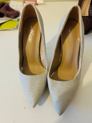 Glittriga silverfärgade pumps från Mustang - Snygga pumps från Mustang i silverfärgat tyg med diskret glittrig yta. Skorna har spetsig tå och höga smala klackar som ger en elegant look. Perfekta för dig som vill sticka ut lite extra. Insidan är beige och materialet känns bekvämt.