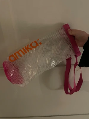 Transparent shoppingväska från Amika - Säljer en transparent väska från Amika med rosa detaljer och orange logga. Väskan har justerbar axelrem i rosa och stängs med ett spänne upptill. Tillverkad i plastmaterial, perfekt för att bära med sig till stranden. Snygg och unik design!