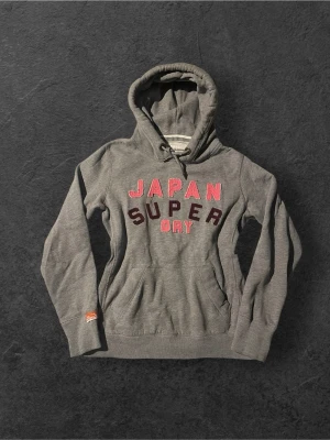 Grå Superdry hoodie med tryck | 40 - Snygg grå hoodie från Superdry med huva och dragsko. Framtill står det 'JAPAN SUPER DRY' i rosa och vinröda bokstäver. Hoodien har en stor magficka och ribbade muddar vid ärmslut och nederkant. Perfekt för en avslappnad streetwear-look.
