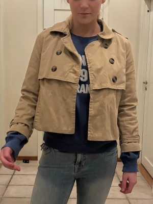 Kort beige trenchcoat med dubbelknäppning - Stilren kort trenchcoat i beige med klassisk dubbelknäppning och axelklaffar. Jackan har krage, dekorativa knappar och ärmar med slejfdetaljer. Perfekt för en trendig och avslappnad look.