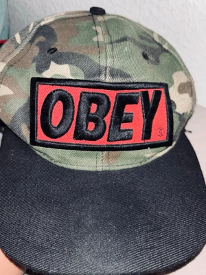 Camo keps från OBEY med logga - Säljer en camouflagemönstrad keps från OBEY med stor broderad logga på framsidan i svart och rött. Skärmen är svart och kepsen har klassisk snapback-modell. Insidan har OBEY-tryck på banden. Perfekt för streetwear-stilen.pris kan diskuteras vid snabbt köp 