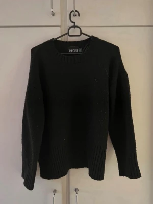 Svart stickad tröja från Pieces - Säljer en svart stickad tröja från Pieces med rund halsringning och ribbade muddar vid ärmslut och nederkant. Tröjan har en avslappnad passform och är perfekt för lager på lager under kyliga dagar.