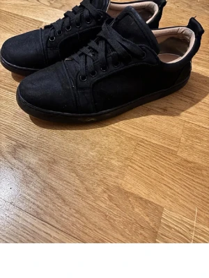 Svarta sneakers med röd sula - Stilrena svarta sneakers med låg profil och röd sula. Skorna har klassisk snörning och ovandel i tygmaterial. Passar perfekt till både jeans och mjukisbyxor för en avslappnad look. Strl 39/40 mycket bra skick dem är bara lite slitna vid snörrerna (se bild) och sulan är lite sliten 