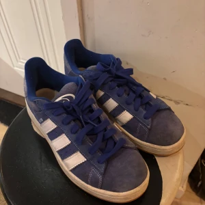 Blå sneakers från Adidas - Snygga blå sneakers från Adidas med vita detaljer och klassiska ränder på sidan. Skorna har snörning och en bekväm sula, perfekt för vardagsbruk.