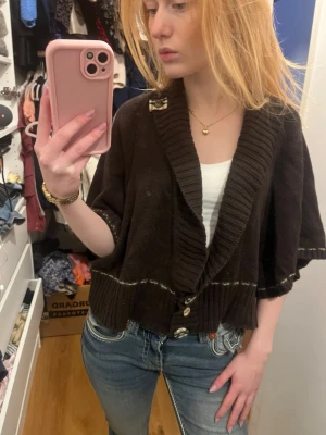 Brun kofta med knappar och krage - Mysig brun kofta med bred ribbad krage och knappar framtill. Modellen är kortare och har vida, halvlånga ärmar. Stickad i mjukt material och har en dekorativ söm i midjan. Perfekt att slänga över en topp för en avslappnad look. Känns inte som L