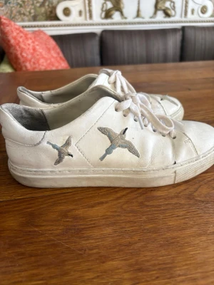 Vita sneakers från ARIGATO med fågelmotiv - Snygga vita sneakers från ARIGATO i läder med broderade fåglar på sidan. Klassisk låg modell med vita snören och diskret logga på hälen. Perfekta för dig som gillar stilrena skor med en unik detalj.  Köpta för 2.300, eftersom defekter förekommer så går priset absolut att diskuteras!