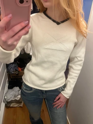 Vit stickad tröja Sea Adventure - Vit stickad tröja från Sea Adventure med svart kant runt v-ringningen och ett diskret geometriskt mönster framtill. Tröjan har lång ärm och är gjord i ett mjukt, tjockare material som passar perfekt till kalla dagar.