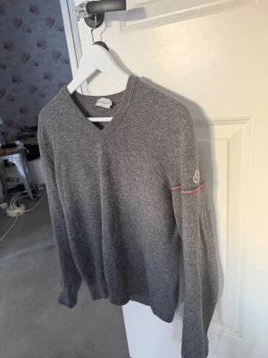 Moncler pull en laine stickad  - Mysig grå stickad tröja från Moncler med V-ringning och diskret Moncler-logga på ärmen. Tröjan har långa ärmar och en tunn röd randdetalj på ärmen. Perfekt för lager på lager och enkel att matcha till olika outfits.