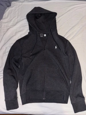 Mörkgrå hoodie från Polo Ralph Lauren - Säljer denna mörkgråa Polo Ralph Lauren zip hoodien eftersom den är för liten för mig. Använd ett fåtal gånger och har inga skador. Köpt på Polo Ralph Lauren för 2295kr. 