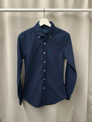 Ralph Lauren Skjorta - En klassisk mörkblå buttondown skjorta ifrån Ralph lauren i storlek XS. Skjortan är använd men svårt att hitta några defekter. Hör gärna av dig vid frågor och funderingar🙌🏼