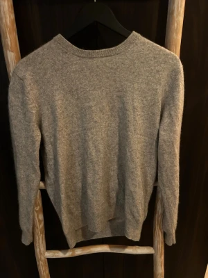 Grå cashmere tröja - Mysig grå stickad tröja med rund hals och långa ärmar. Tröjan har en enkel, klassisk design och är tillverkad i mjuk ullmix som passar perfekt för kyligare dagar. Ribbstickade muddar vid ärmslut och nederkant ger en snygg finish.