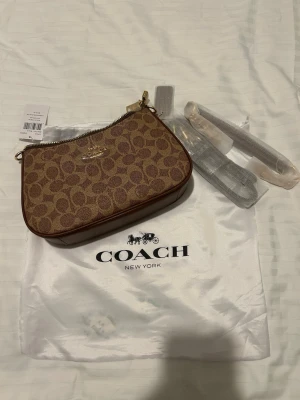 Coach brun monogram handväska - Snygg handväska från Coach i brunt monogramtyg med klassiskt C-mönster och guldfärgade detaljer. Väskan har dragkedja upptill, innerfack och är fodrad i matchande brunt tyg. Perfekt storlek för det viktigaste och levereras med dustbag.