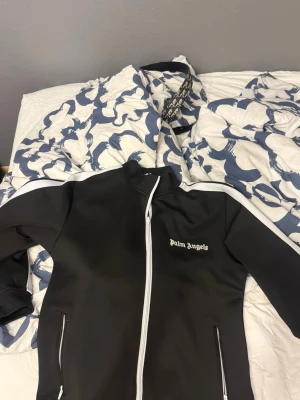 Svart ziptröja från Palm Angels - Säljer en svart ziptröja från Palm Angels med vit dragkedja och vita detaljer längs axlarna. Tröjan har hög krage, två fickor med dragkedja och logga på bröstet. Perfekt för en sportig och trendig look. Priset är ej fast