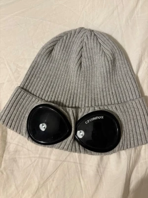 Grå ribbstickad mössa C.P. Company - Säljer en grå ribbstickad mössa från C.P. Company med ikoniska svarta glasögonlinser på uppviket. Mössan är tillverkad i mjuk ull och har en modern, streetinspirerad look som sticker ut. Perfekt accessoar för dig som gillar unik och trendig stil.