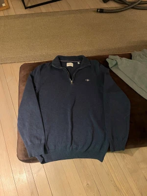 Mörkblå stickad tröja från GANT - Snygg mörkblå stickad tröja från GANT med halv dragkedja och ribbad krage. Tröjan har långa ärmar, ribbade muddar och en liten GANT-logga broderad på bröstet. Perfekt för lager på lager och klassisk stil.