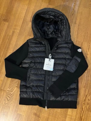 Svart pufferjacka från Moncler - Snygg svart pufferjacka från Moncler med stickade ärmar och huva. Jackan har dragkedja framtill, ribbade muddar och en Moncler-logga på ärmen. Perfekt för kyliga dagar och ger en sportig men stilren look.