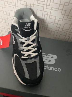 New Balance 530 svarta sneakers - Snygga New Balance 530 sneakers i svart, grått och vitt med klassisk mesh och mocka. Skorna har vita snören, tydlig logga på sidan och bekväm sula med ABZORB-dämpning. Perfekta för dig som gillar sportig och stilren look.