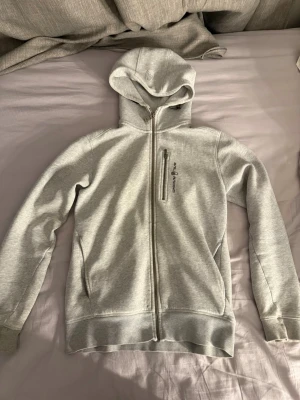 Ljusgrå hoodie från Sail racing  - Ljusgrå hoodie med dragkedja från Sail racing. Tröjan har en huva, två sidofickor och en bröstficka med dragkedja och logga. Tillverkad i mjukt sweatshirtmaterial, perfekt för chill dagar. Klassisk passform och ribbade muddar vid ärmar och nederkant.