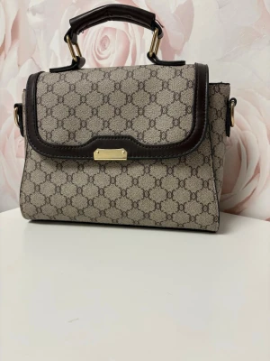 Beige handväska med monogram och läderdetaljer - Snygg beige handväska  med klassiskt monogrammönster och mörkbruna läderdetaljer. Väskan har ett topphandtag med guldfärgade metalldetaljer och en elegant rektangulär form. Perfekt för dig som gillar tidlös design och exklusiva accessoarer. Om du vill ha fler bilder inga problem det är bara att skicka och be. Aldrig använd!