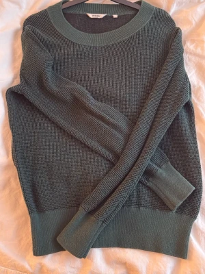 Mörkgrön stickad tröja  - Mysig mörkgrön stickad tröja från Wrangler med rund halsringning och breda ribbade muddar vid ärmslut och nederkant. Tröjan har en klassisk, avslappnad passform och är perfekt för lager på lager under kyliga dagar.