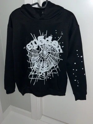 Svart hoodie med spindelnät-tryck - Cool svart hoodie från Snigeln med stort vitt spindelnät och text på bröstet samt små stjärnor på ena ärmen. Klassisk huva och ribbade muddar. Perfekt för dig som gillar streetwear och vill sticka ut med en unik design.