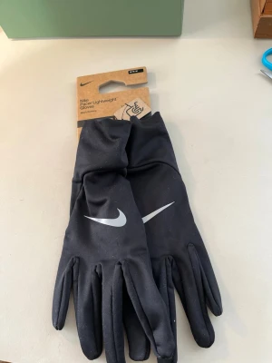 Nike svarta Therma-Fit löparhandskar - Svarta Nike Pacer Lightweight löparhandskar med Therma-Fit-teknologi. Handskarna har en stilren design med vit Nike-logga på ovansidan och är tunna och smidiga för löpning eller träning utomhus.