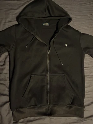 Svart hoodie från Polo Ralph Lauren - Klassisk svart hoodie med dragkedja från Polo Ralph Lauren. Har huva med dragsko, två stora fickor framtill och den ikoniska vita logon broderad på bröstet. Tillverkad i mjukt bomullsmaterial som är skönt mot huden. Perfekt för en avslappnad och stilren look.