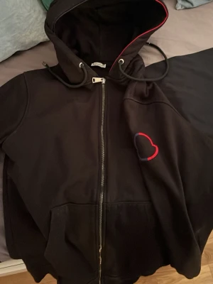 Svart Moncler hoodie med dragkedja - Svart hoodie från Moncler med hel dragkedja, huva med dragsko och röd detaljsöm på insidan av huvan. Bröstet pryds av en broderad Moncler-logga i rött och blått. Praktiska kängurufickor framtill. Perfekt för en stilren och sportig look.
