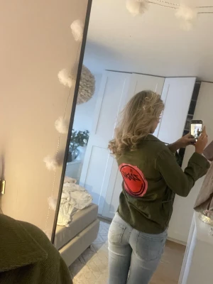 Grön overshirt från Zadig Voltaire  - Säljer en grön overshirt från Zadig Voltaire med en stor röd logga på ryggen. Jackan har klassisk krage, knäppning framtill och fickor på sidorna. Perfekt för dig som gillar streetwear och Stockholmstil och vill sticka ut lite extra.