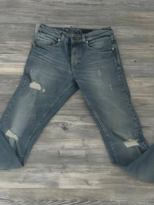 Ljus blå Tiger Of Sweden jeans me lite slitningar  - Ett par blå jeans med slitningar och hål på båda benen. Klassisk femficksmodell med normal passform och dragkedjegylf. Jeansen har en tvättad look och detaljerade slitningar för en trendig, avslappnad stil.