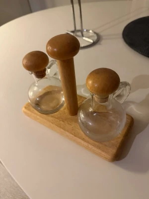 Oljeflaskor i glas med träställ - Två små runda oljeflaskor i klart glas med träkorkar och handtag, placerade i ett stilrent ställ av ljust trä. Perfekt för olja och vinäger på matbordet. Enkel och snygg design som passar i de flesta kök.             