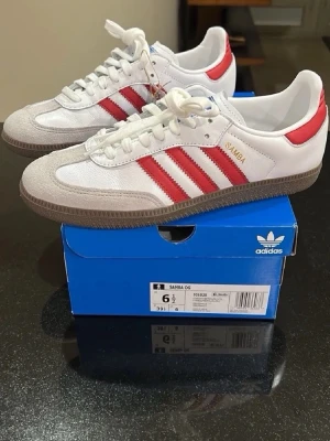 Adidas Samba - Klassiska Adidas Samba OG sneakers i vitt läder med röda detaljer och de ikoniska tre ränderna på sidan. Skorna har grå mockadetaljer vid tån och en brun gummisula. Perfekt retrostil med snörning och låg profil.