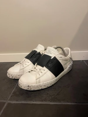 Valentino vita sneakers med svart band - Snygga vita sneakers från Valentino med ett brett svart band över vristen och svartprickig sula. Skorna har klassisk låg profil, vita skosnören och diskret logga på plösen. Tillverkade i läder för en clean och exklusiv look.