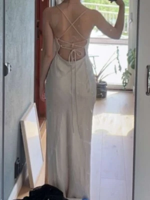 Lång champagnefärgad klänning med snörning - Elegant långklänning i champagnefärg med smala axelband och öppen rygg med snygg snörningsdetalj. Klänningen har en figurnära passform och är tillverkad i silkes matreal. Jättefin balklänning för 9an.