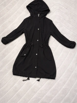 Svart lång vindjacka med huva - Svart tunjacka med huva och dragsko i midjan samt nederkant. Jackan har tryckknappar framtill, två stora fickor med lock och långa ärmar. Perfekt för sommarens dagar och enkel att matcha till olika outfits.
