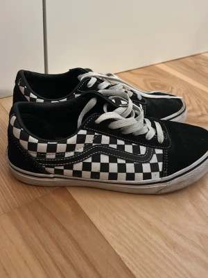 Vans Old Skool Checkerboard sneakers - Klassiska Vans Old Skool sneakers med svart och vitt checkerboard-mönster på sidan, svart mocka framtill och vita snören.  Perfekta för en streetwear-look och passar till det mesta.