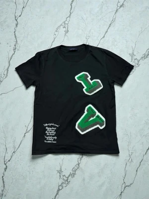 Svart t-shirt med gröna bokstäver - Cool svart t-shirt med stora gröna och grå bokstäver i frotté på framsidan. T-shirten har rund hals och korta ärmar. Det finns även vit text broderad på ena sidan nedtill. Perfekt för dig som gillar streetwear och unika detaljer.