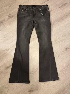 Grå bootcut jeans från True Religion - Säljer ett par snygga grå bootcut jeans från True Religion. Jeansen har klassisk femficksmodell, silverfärgade nitar och etikett i midjan. Materialet är mjukt denim med lätt tvättad look och utsvängda ben. Perfekta för en trendig och avslappnad stil.