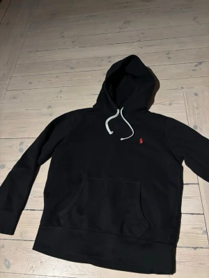 Svart hoodie från Polo Ralph Lauren - Svart hoodie från Polo Ralph Lauren med vit dragsko och klassisk röd logga broderad på bröstet. Hoodien har känguruficka framtill och är tillverkad i mjukt bomullsmaterial. Perfekt för en avslappnad och stilren look.